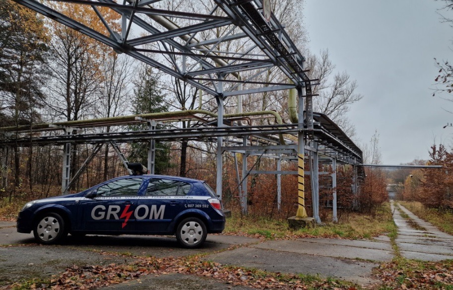 Granatowy samochód z napisem 'GROM' na tle metalowej konstrukcji przemysłowej i drzew bez liści, jesienna sceneria.