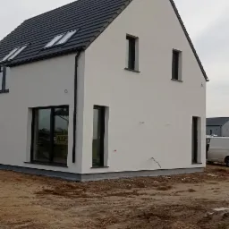 Nowoczesny dom jednorodzinny z białymi ścianami i ciemną dachówką, widok z zewnątrz na niezagospodarowaną działkę.