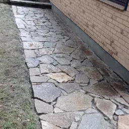 Ścieżka z nieregularnych kamieni prowadząca wzdłuż ceglanego budynku, widoczne schody i złożone krzesło turystyczne.