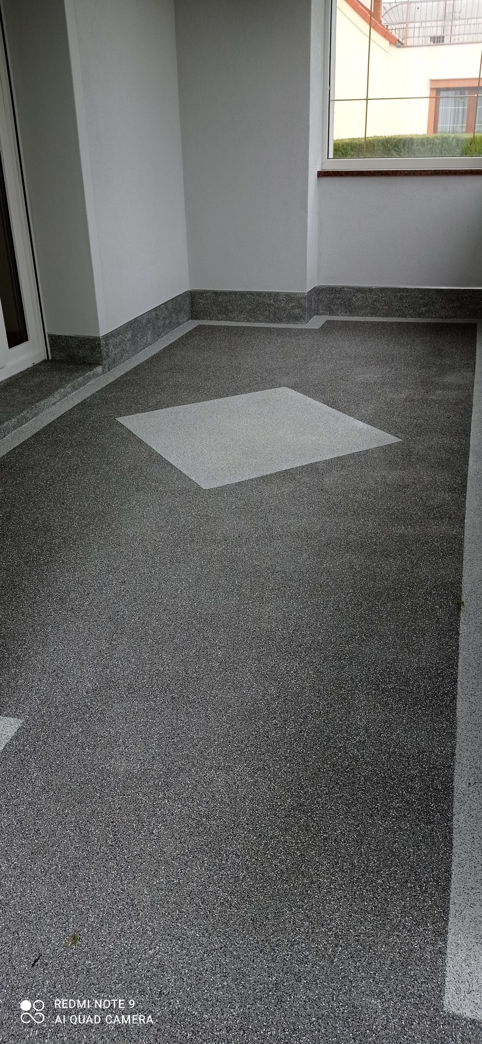 Szary taras z żywicy epoksydowej z dekoracyjnym, jasnoszarym kwadratem w centralnej części, wykończony szarym cokołem przy ścianach.