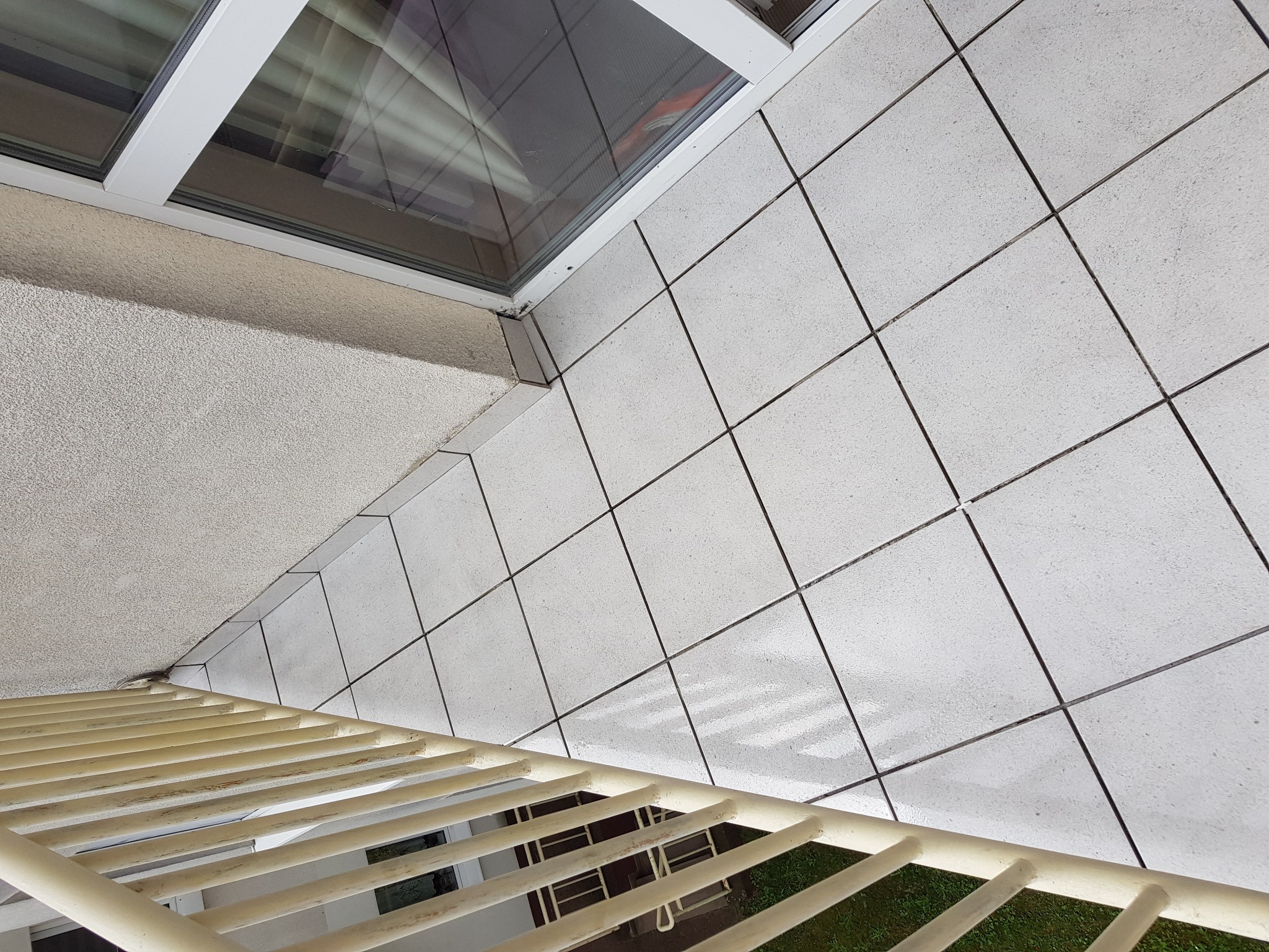 Białe płytki na balkonie widziane z góry, z widoczną barierką i fragmentem okna, z odbiciem światła na powierzchni płytek.