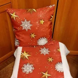 Czerwona poduszka i bieżnik z motywem śnieżynek i gwiazd, ułożone na białej pościeli, na tle drewnianej szafki.