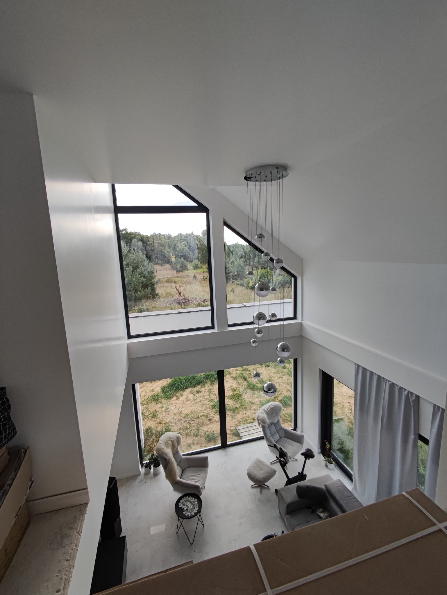 Wysoki salon z dużymi oknami wychodzącymi na zieleń, nowoczesny żyrandol z kul, szare meble i białe ściany, widok z góry.