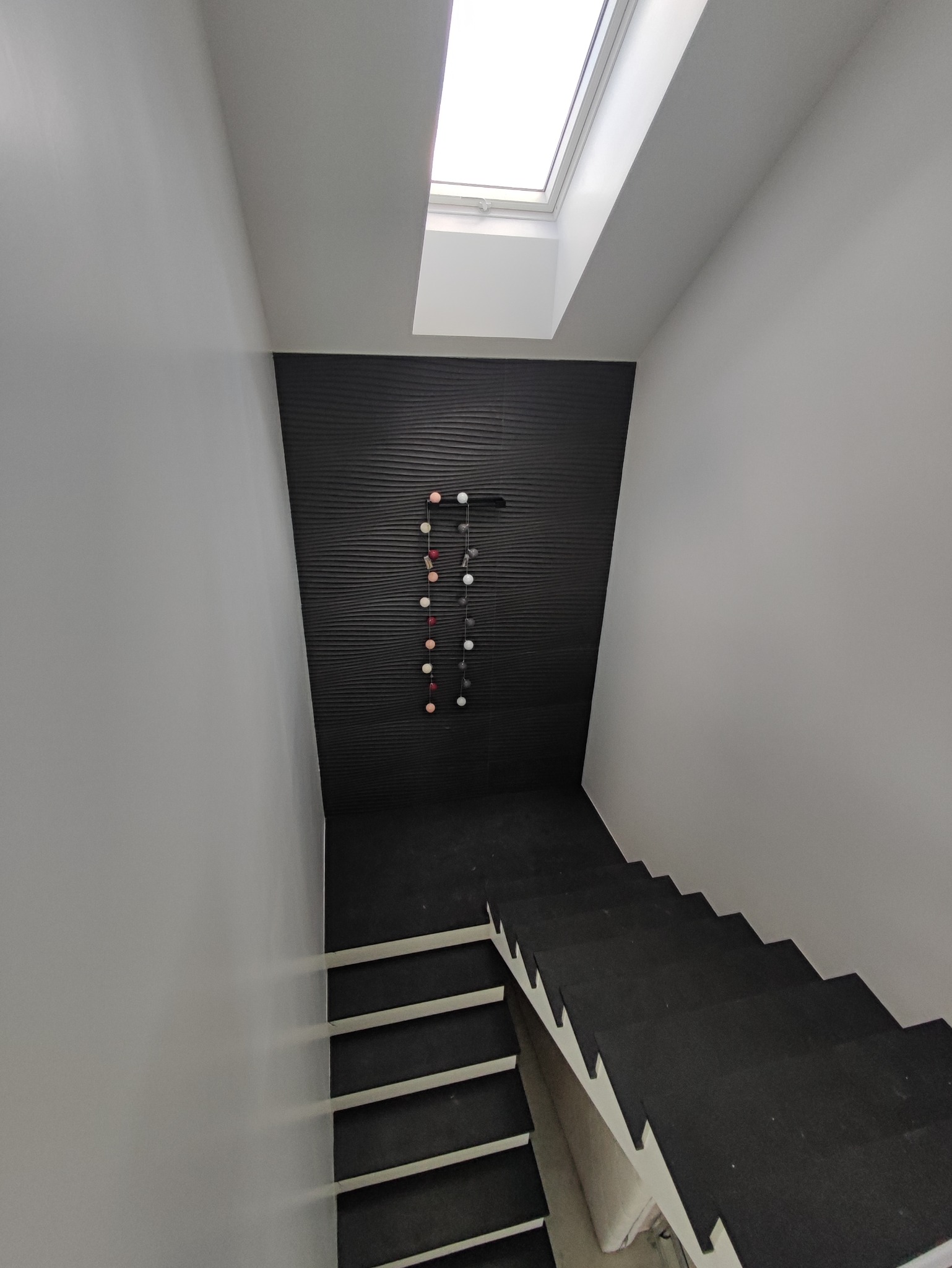 Nowoczesne schody w dół z czarnymi stopniami i białymi podstopnicami, czarna, teksturowana ściana z dekoracją w postaci girlandy świetlnej i okno dachowe w wąskim korytarzu.