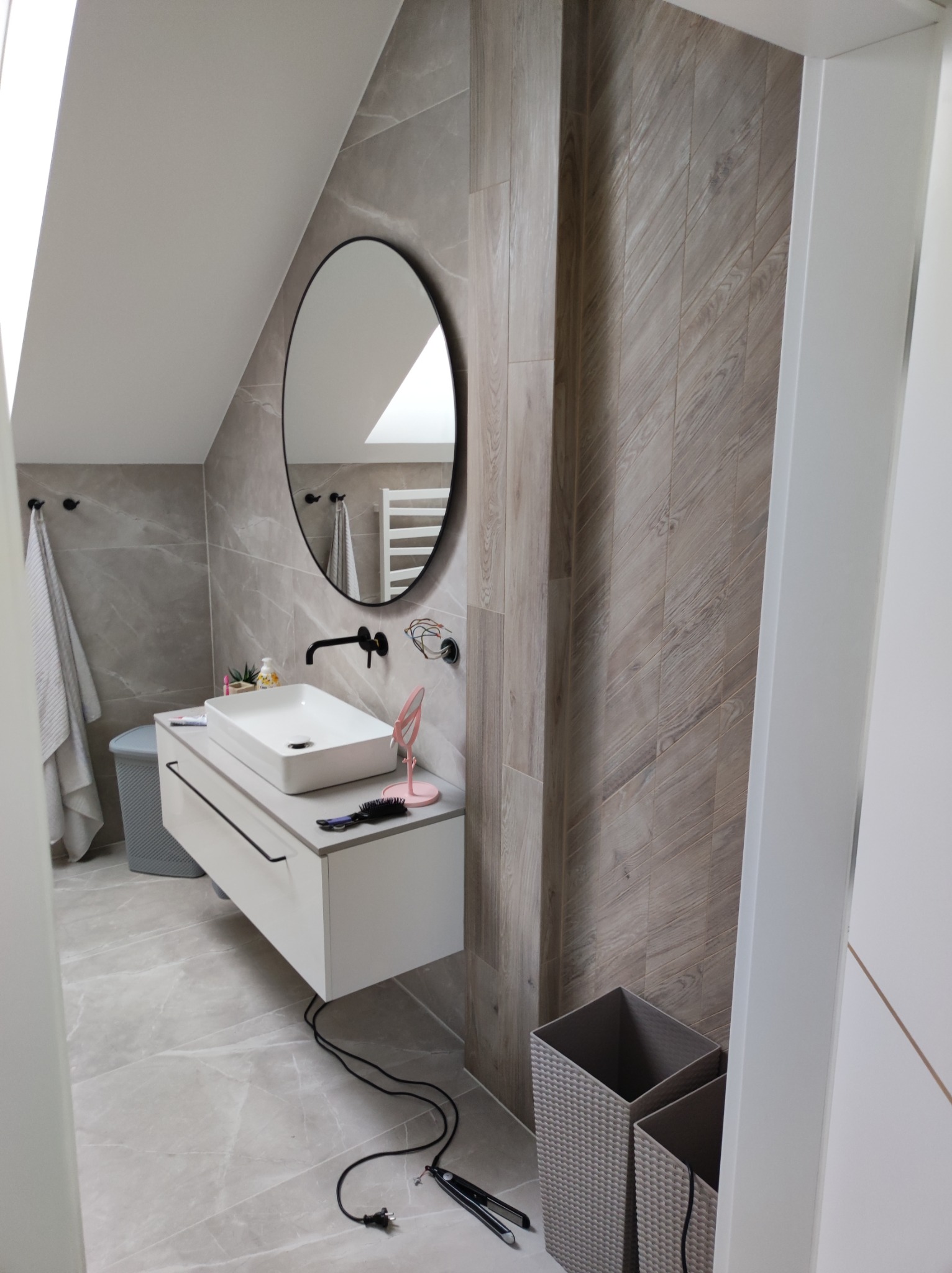 Nowoczesna łazienka na poddaszu z marmurowymi płytkami, owalnym lustrem w czarnej ramie, białą umywalką nablatową i drewnopodobną ścianą z ukośnym wzorem; na podłodze leży prostownica do włosów.