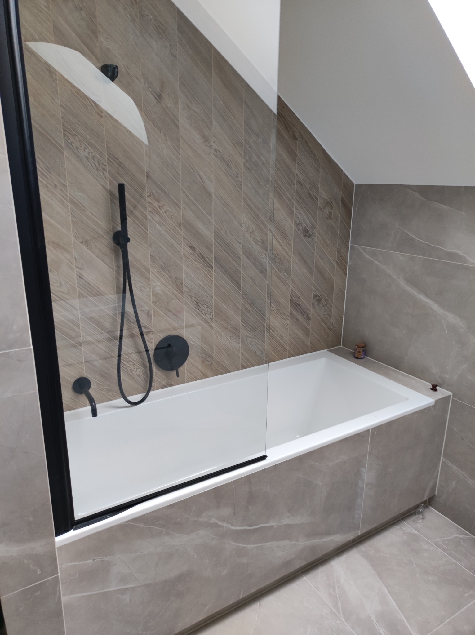 Wykończona łazienka na poddaszu z wanną obudowaną płytkami imitującymi marmur, przeszklonym parawanem nawannowym z czarną ramą i czarną armaturą.