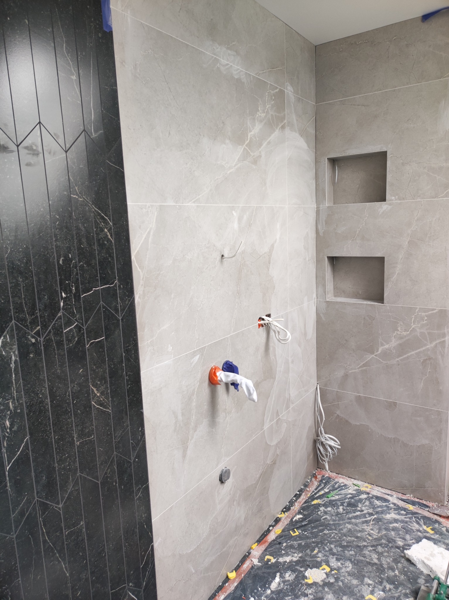 Ściana łazienki w trakcie wykańczania, widoczne płytki imitujące marmur w odcieniach szarości, czarne płytki ułożone we wzór jodełki, puszki elektryczne z wystającymi kablami, wnęki w ścianie...