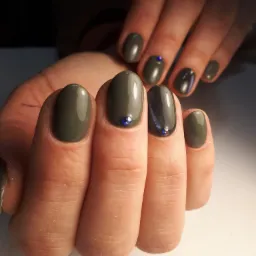 Dłonie z oliwkowymi paznokciami o zaokrąglonym kształcie, ozdobione pojedynczymi, drobnymi, niebieskimi kryształkami. Jeden paznokieć w kolorze ciemnego grafitu z efektem kociego oka.