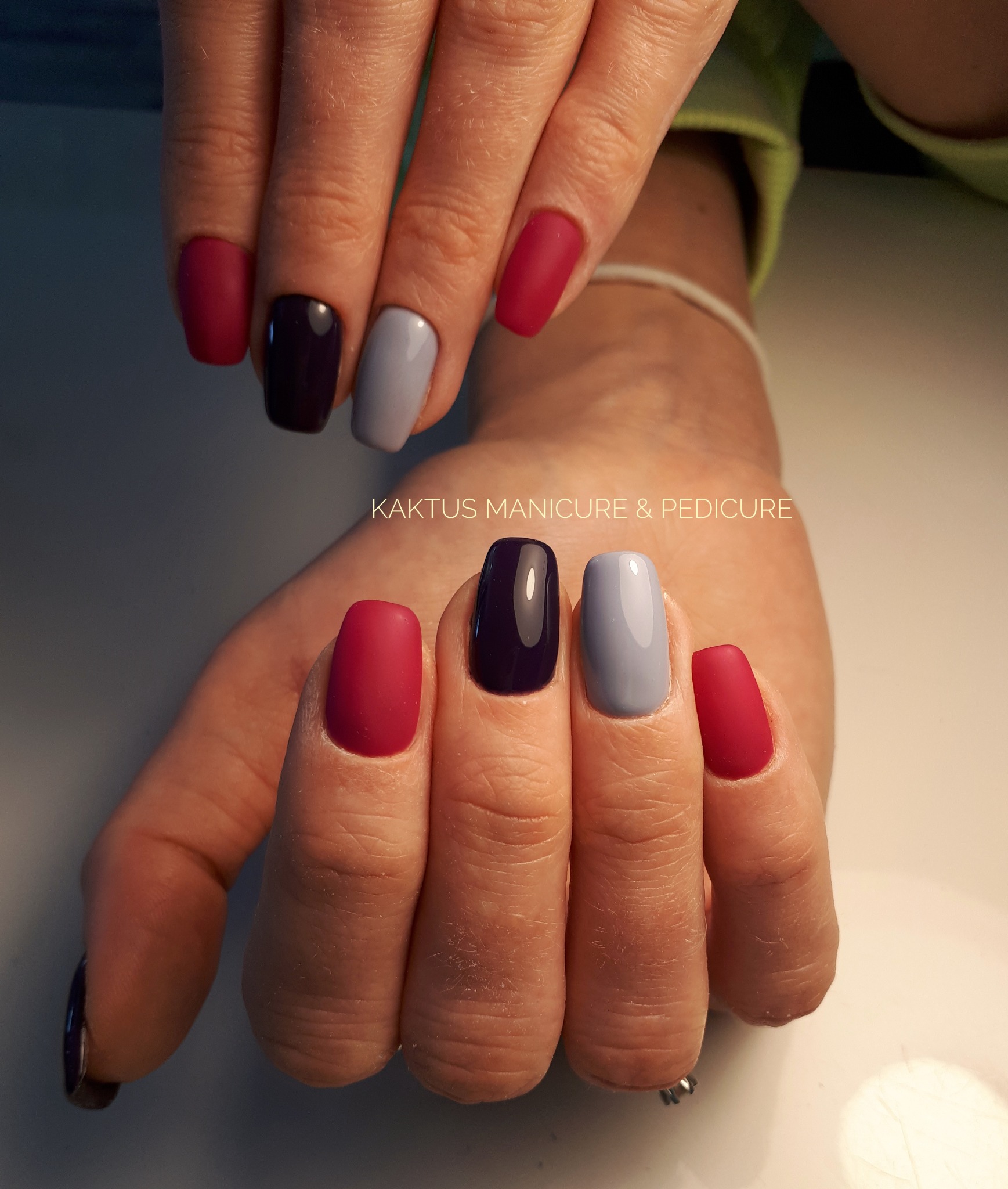 Dłonie z pomalowanymi paznokciami w kolorach: matowy czerwony, błyszczący ciemnofioletowy i jasnoszary, na tle jasnego blatu, w tle widoczne logo 'Kaktus Manicure & Pedicure'.