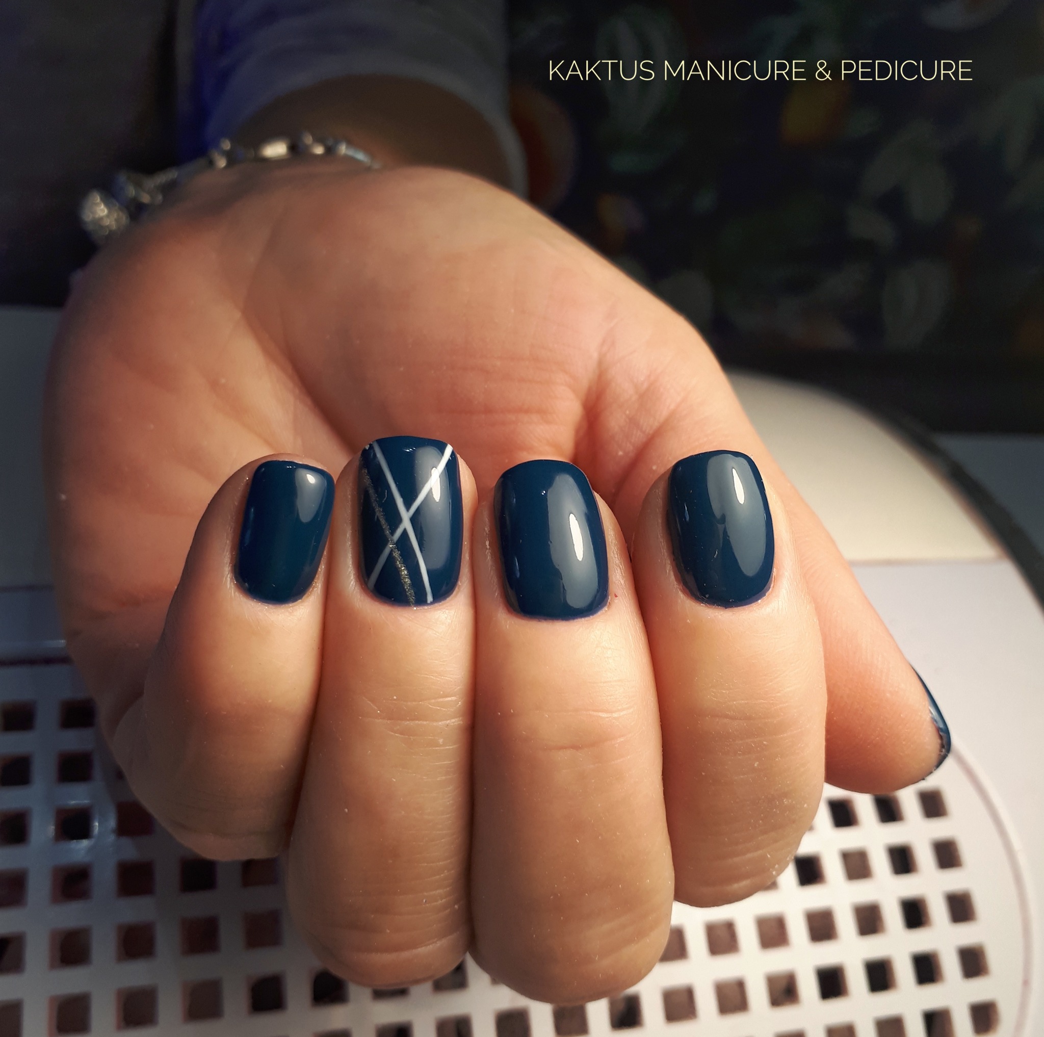 Dłoń z granatowymi, błyszczącymi paznokciami; jeden paznokieć ozdobiony białymi i złotymi liniami na krzyż.