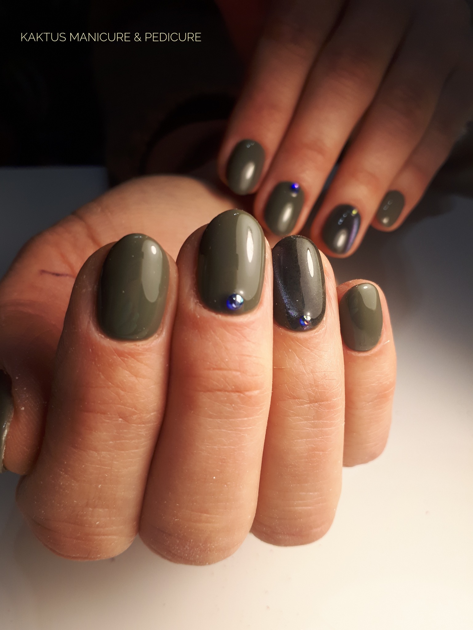 Dłonie z oliwkowymi paznokciami o zaokrąglonym kształcie, ozdobione pojedynczymi, drobnymi, niebieskimi kryształkami. Jeden paznokieć w kolorze ciemnego grafitu z efektem kociego oka.