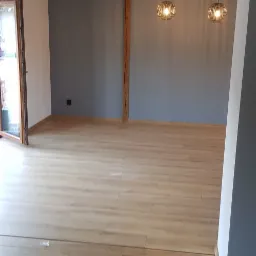 Puste, odświeżone wnętrze z drewnianą belką stropową, szarą ścianą, parkietem w odcieniach drewna i dwoma wiszącymi lampami w stylu industrialnym.