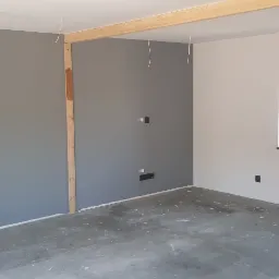 Pokój po częściowym malowaniu ścian na szaro i biało, widoczne drewniane elementy konstrukcyjne, kable wystające z sufitu oraz ślady po pracach na podłodze.