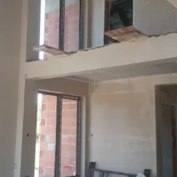 Surowe wnętrze dwukondygnacyjnego domu w trakcie prac wykończeniowych, ściany pokryte wstępną warstwą tynku, widoczne okna z ceglanymi ścianami w tle, przewody elektryczne wystające ze ścian...