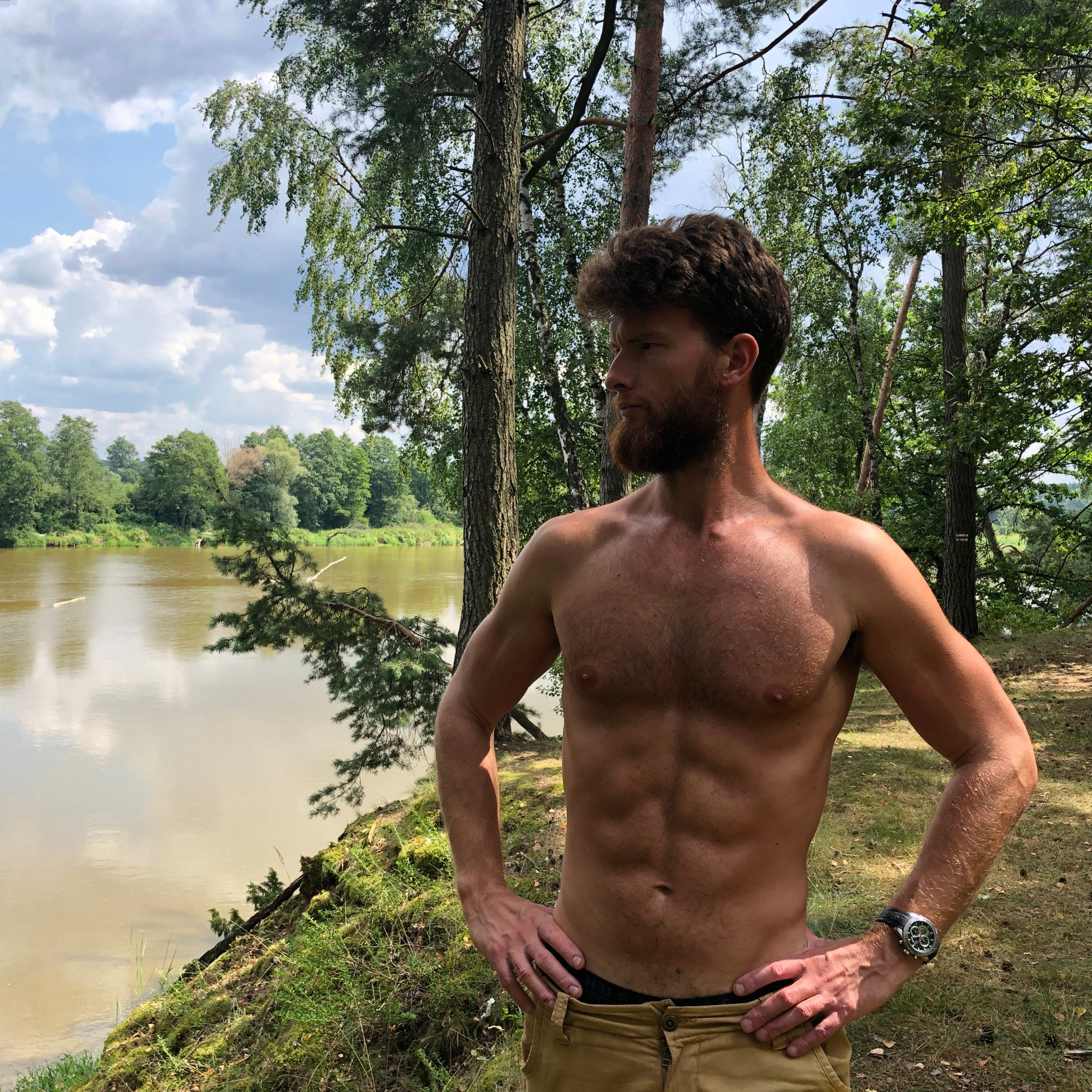 Mężczyzna z brodą, bez koszulki, prezentuje wyrzeźbioną sylwetkę na tle rzeki i drzew.