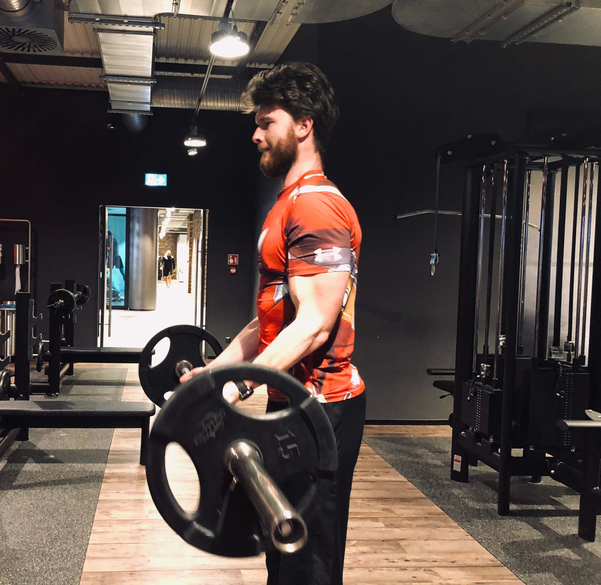 Mężczyzna z brodą w sportowej koszulce wykonuje ćwiczenie bicepsów z obciążeniem 15 kg na siłowni, w tle widoczny sprzęt do ćwiczeń i wyjście.