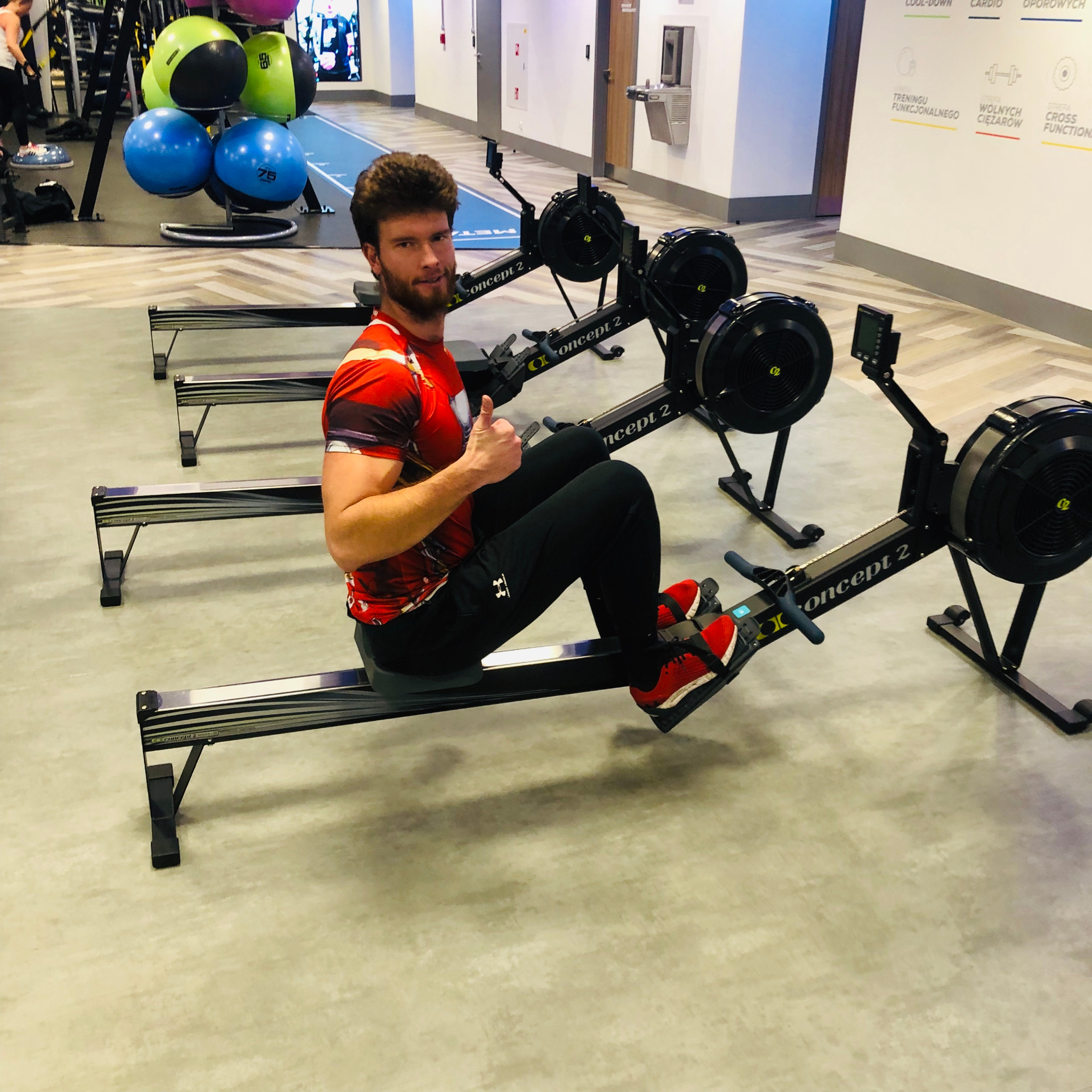 Mężczyzna na ergometrze wioślarskim Concept 2 w siłowni, ubrany w czerwoną koszulkę i czarne spodnie, pokazuje kciuk w górę.