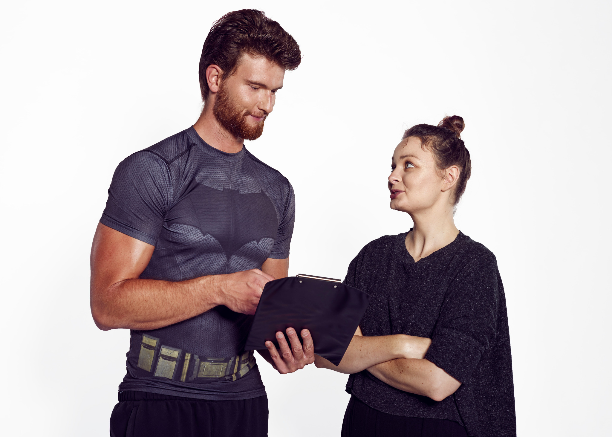 Mężczyzna w koszulce z motywem Batmana trzymający czarną podkładkę z dokumentami rozmawia z kobietą na białym tle.