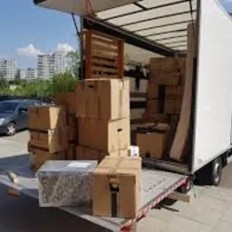 Otwarta paka białej ciężarówki załadowana kartonami i elementami meblowymi podczas przeprowadzki, widoczne budynki w tle.