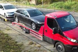 Czerwona laweta z dwoma samochodami osobowymi na platformie, czarnym kombi i białym hatchbackiem, zaparkowana na poboczu drogi w słoneczny dzień.