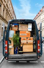 Wnętrze busa załadowane kartonami, rośliną doniczkową i innymi przedmiotami gotowymi do transportu, widoczne otwarte tylne drzwi pojazdu na tle miejskiej ulicy z budynkami.
