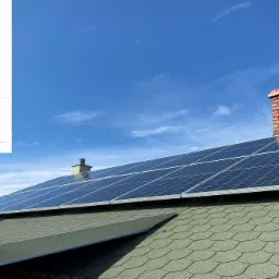 Panele fotowoltaiczne na dachu z zieloną dachówką, komin z czerwonej cegły i wykres produkcji energii na tle błękitnego nieba.