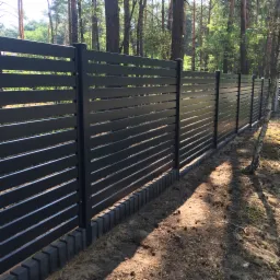 Długi, poziomy, nowoczesny płot z ciemnych, metalowych paneli ułożonych poziomo, z widocznymi przerwami między nimi, osadzony na betonowej podmurówce z kostki, na tle leśnego krajobrazu z wysokimi...