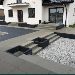 Nowoczesna aranżacja przestrzeni przed domem z wykorzystaniem szarych płyt betonowych i kostki granitowej; schody z donicami wkomponowane w nawierzchnię.