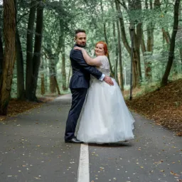 Para młoda, on w granatowym garniturze, ona w białej sukni, pozują na leśnej drodze z białą linią pośrodku, otoczeni drzewami.