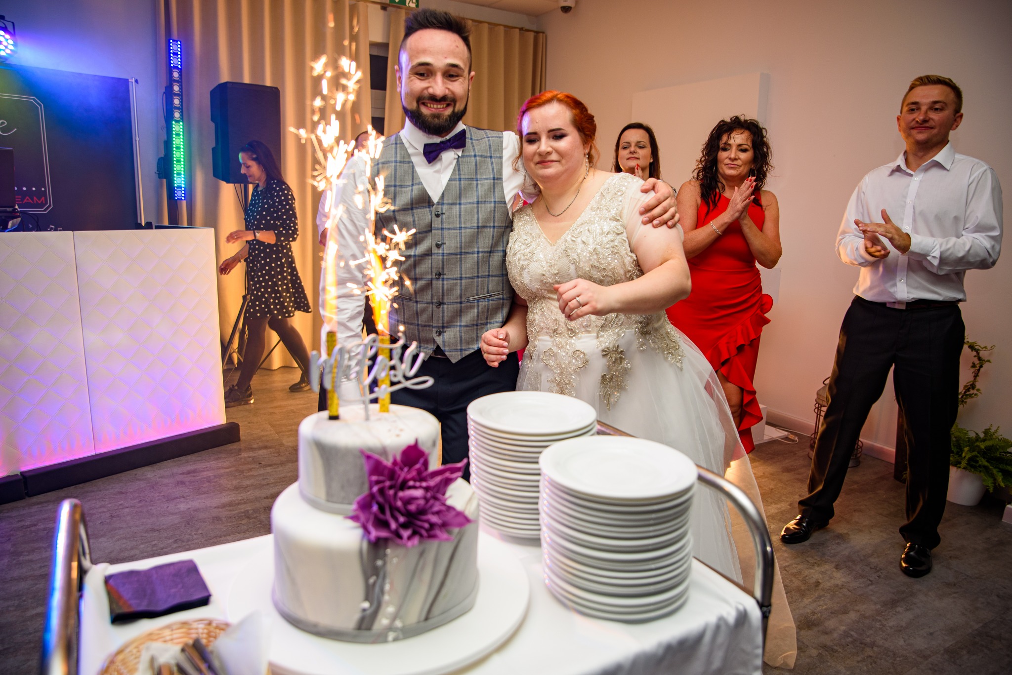 Uśmiechnięta para młoda pozuje z tortem weselnym z fontannami iskier, w tle goście klaszczą, widoczny DJ i stosy talerzy.