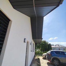 Arkadiusz Halat - Biała elewacja domu jednorodzinnego z szarym podbitką dachową i oknem z kratami. Widoczne oświetlenie zewnętrzne i fragment podjazdu z samochodami w słoneczny dzień.