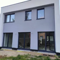 Dwukondygnacyjny dom jednorodzinny w Lublinie z szarą elewacją i czarną stolarką okienną, widoczne ślady prac wykończeniowych wokół budynku.