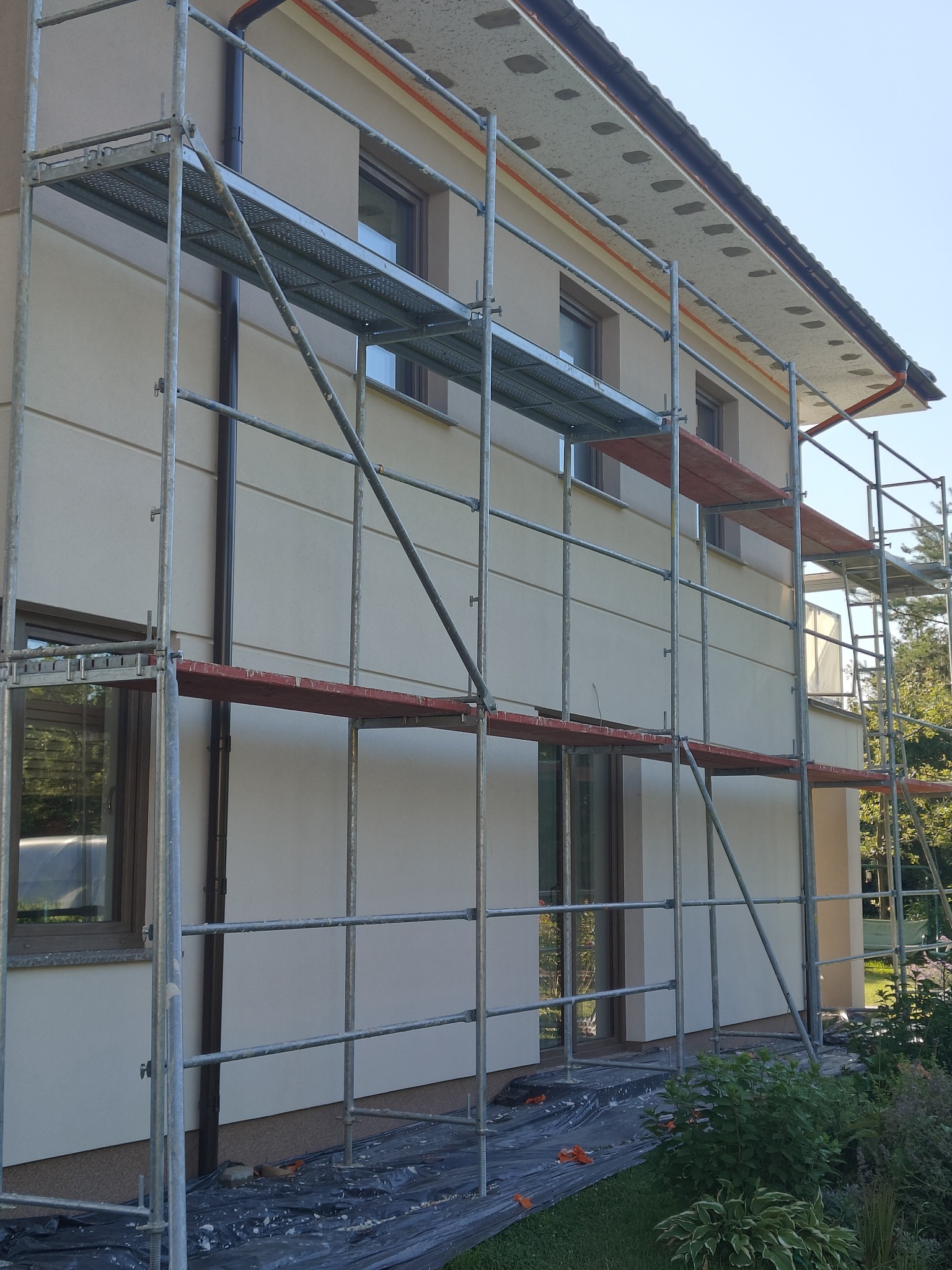 Elewacja domu w trakcie prac z rusztowaniem. Widoczne wykończone fragmenty ściany w jasnym odcieniu. Na dole folia ochronna i roślinność.