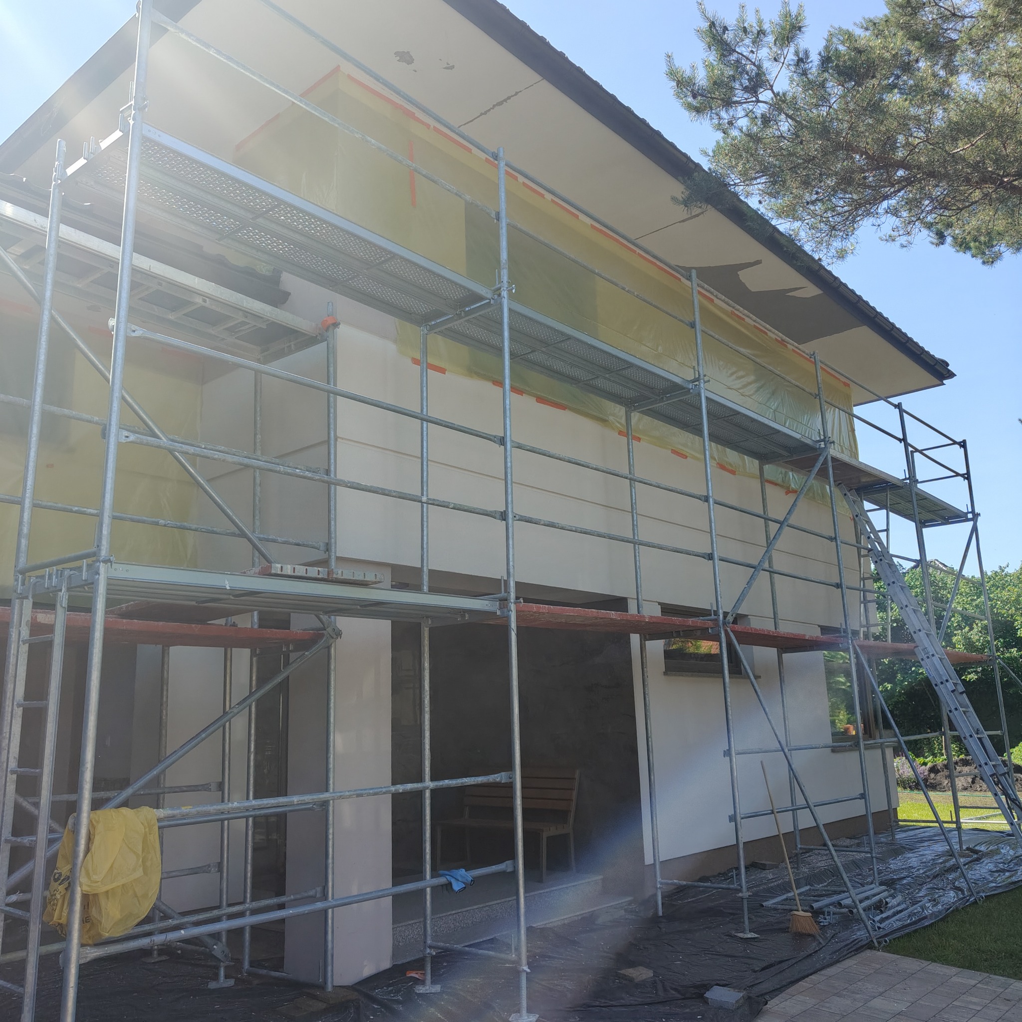 Dom w trakcie prac elewacyjnych, otoczony rusztowaniem. Część elewacji zabezpieczona folią, widoczne narzędzia i materiały budowlane. Jasne, naturalne oświetlenie.