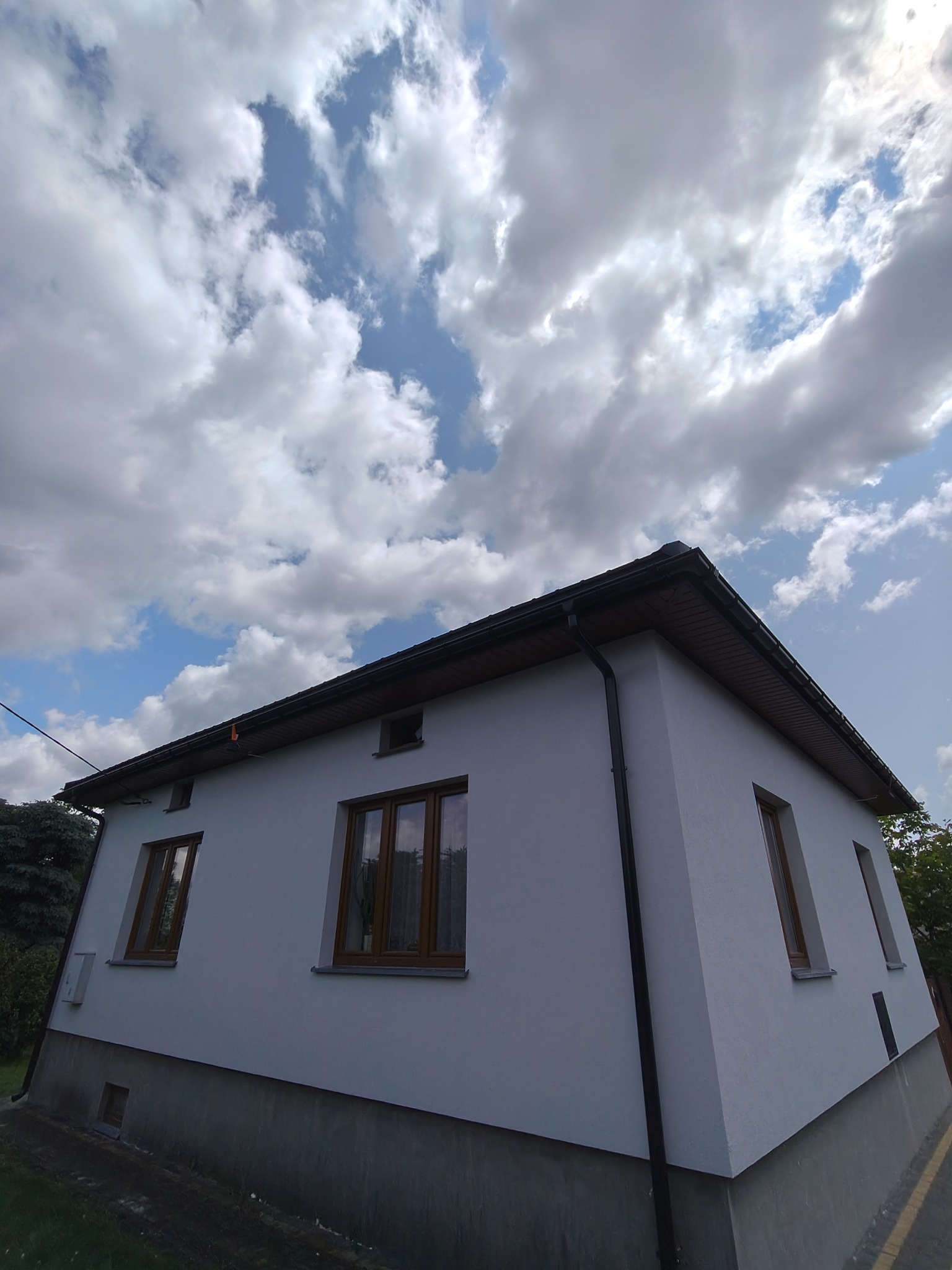 Jasny, otynkowany dom jednorodzinny z brązowymi oknami i ciemnym dachem na tle błękitnego nieba z obłokami. Ujęcie z dołu podkreśla bryłę budynku.
