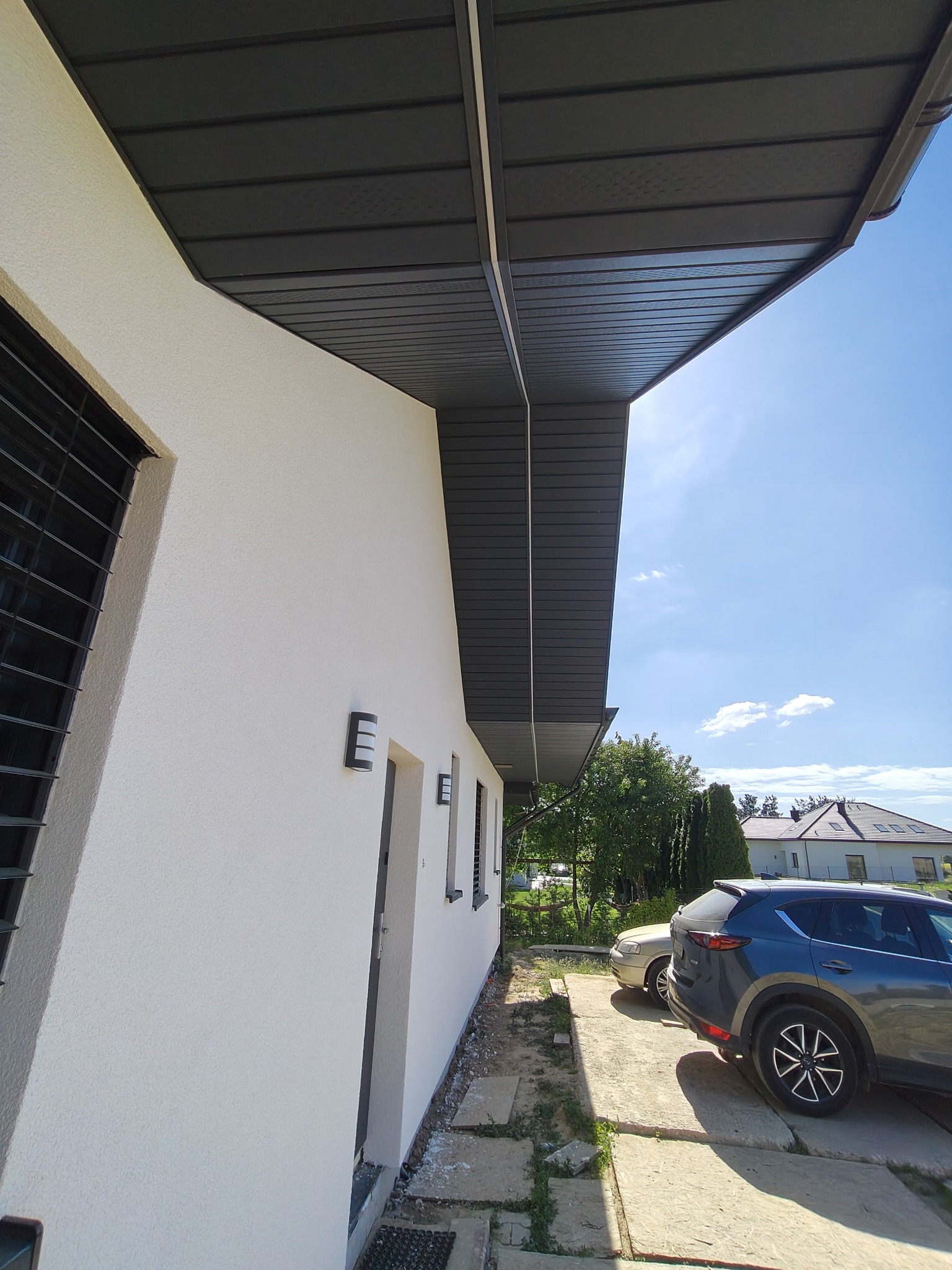 Biała elewacja domu jednorodzinnego z szarym podbitką dachową i oknem z kratami. Widoczne oświetlenie zewnętrzne i fragment podjazdu z samochodami w słoneczny dzień.