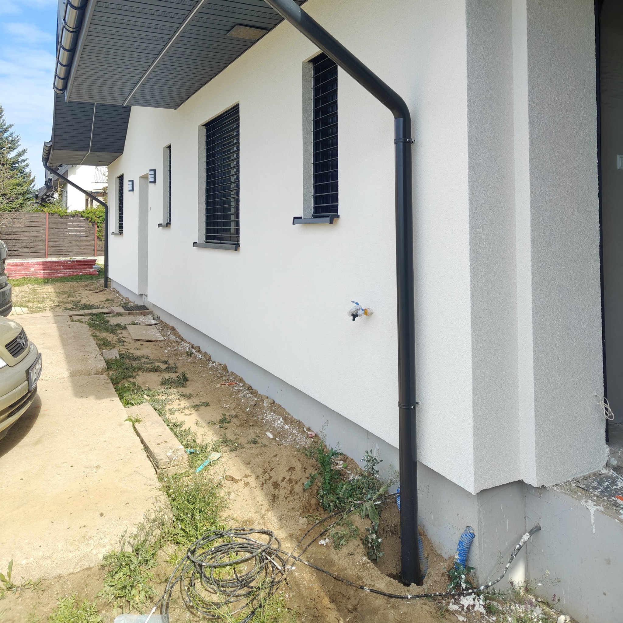 Biała elewacja domu jednorodzinnego z czarnymi oknami i rynnami. Widoczne fragmenty ogrodu i podjazdu. Świeżo położony tynk o wyraźnej fakturze.