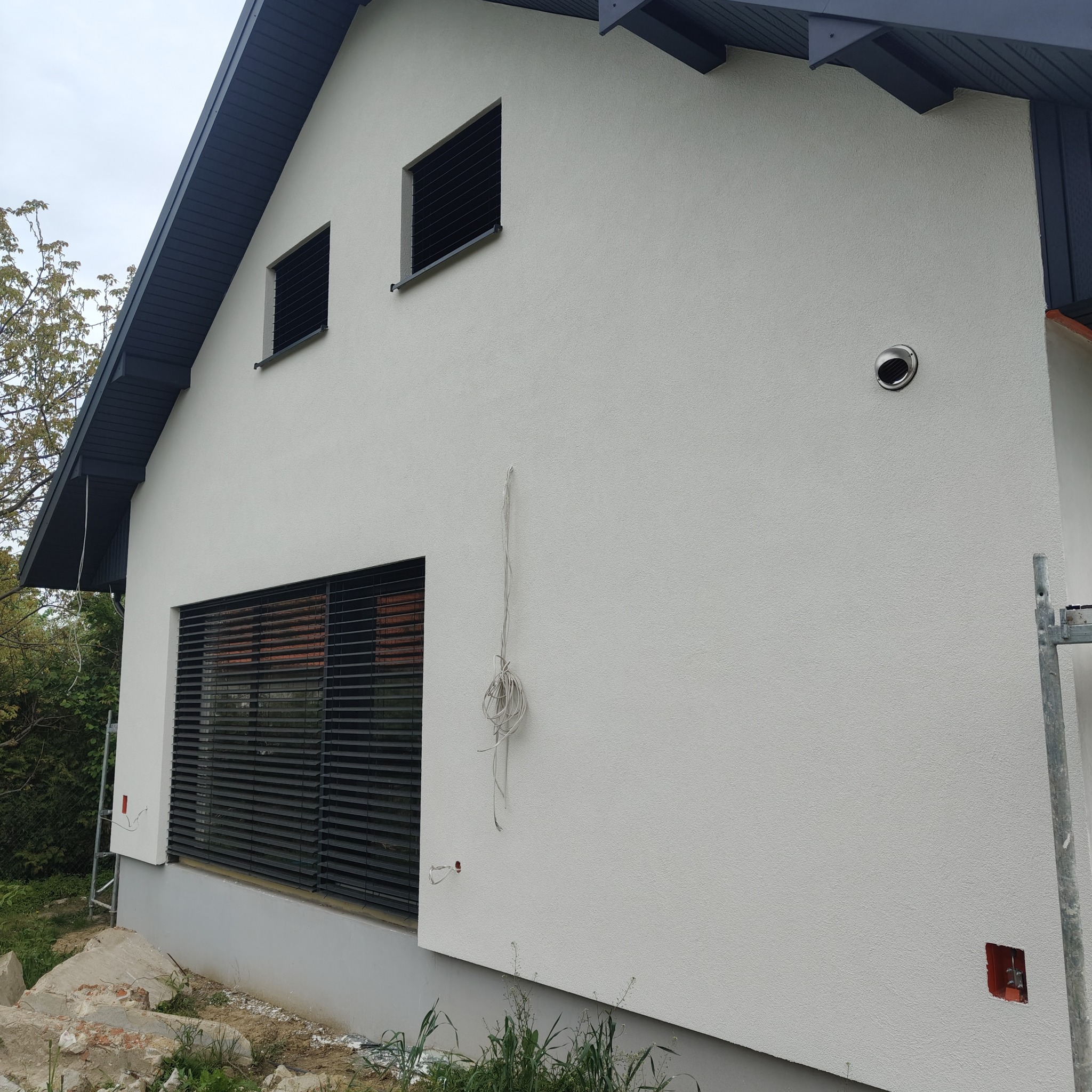 Boczny widok domu po tynkowaniu elewacji na biało, z ciemnymi ramami okiennymi i szarą podbitką dachu. Widoczne kable zwisające ze ściany i fragment rusztowania.