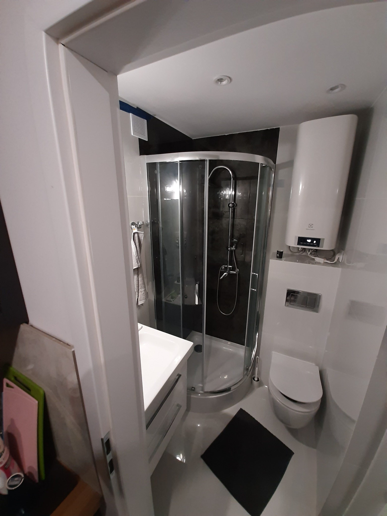 Nowoczesna, jasna łazienka z kabiną prysznicową z zaokrąglonym szkłem, białą umywalką z szafką, podgrzewaczem wody Electrolux i toaletą. Na podłodze czarny dywanik.