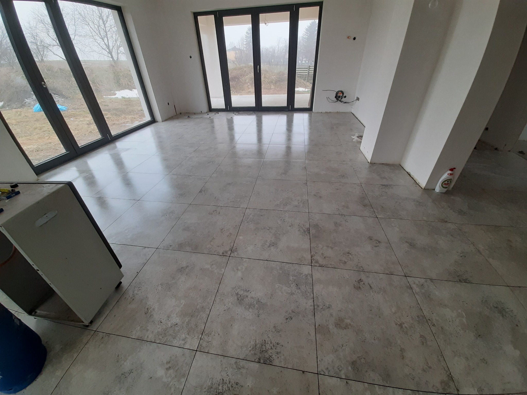 Wnętrze z dużymi oknami i szarymi płytkami podłogowymi, widoczne przygotowania do prac wykończeniowych, butelka płynu do mycia naczyń na podłodze, elementy instalacji elektrycznej wystające ze ściany.