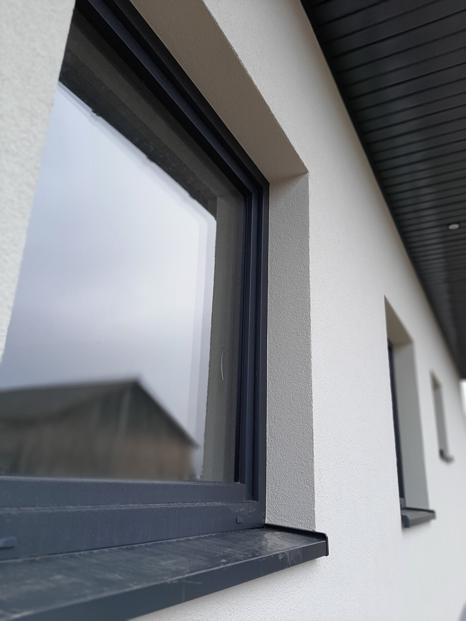 Detal elewacji z oknami o grafitowych ramach, widoczna faktura tynku baranka i fragment podbitki dachowej z ciemnego drewna.