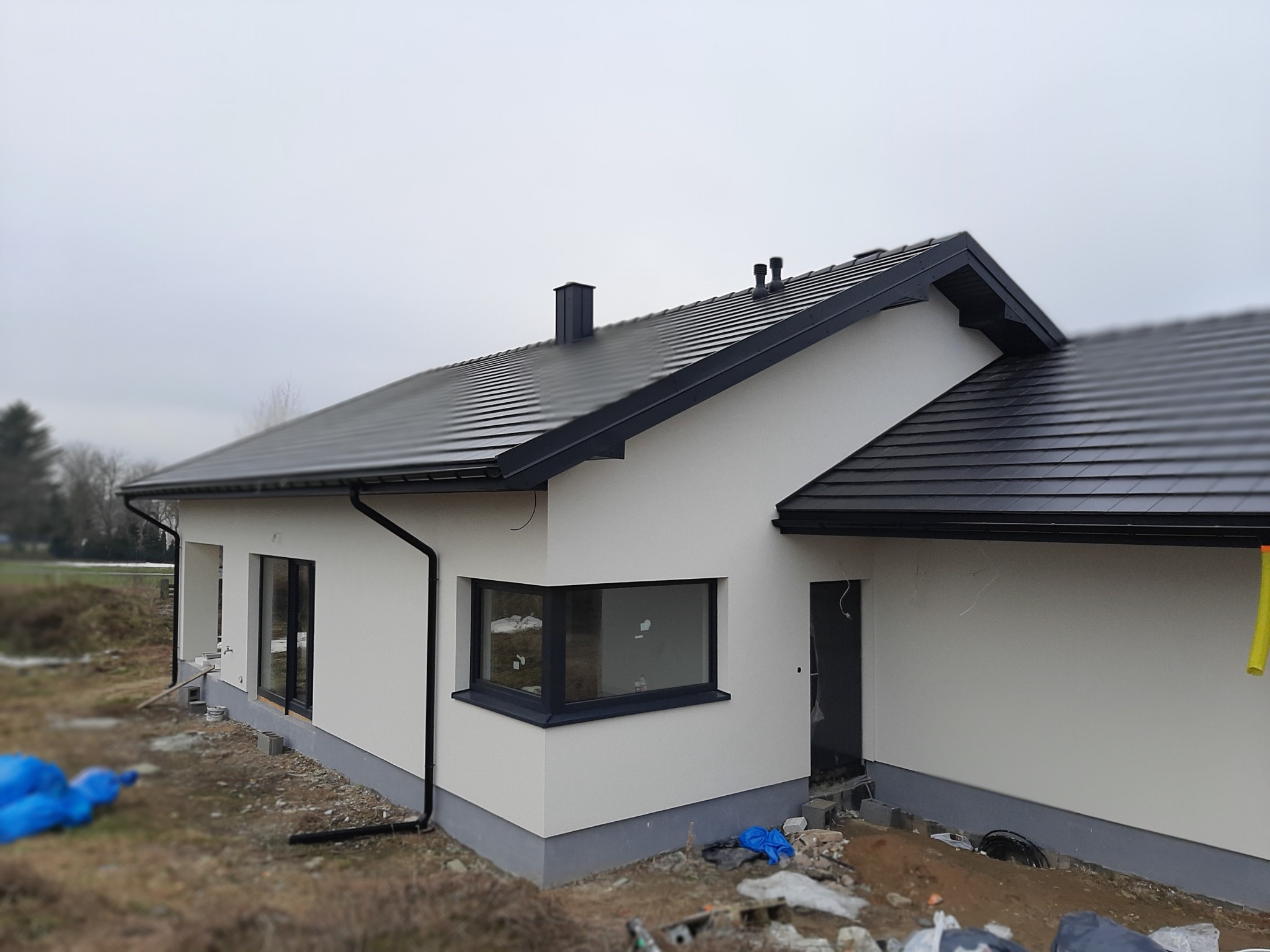 Nowoczesny dom jednorodzinny z ocieploną elewacją w kolorze białym i czarną dachówką, widoczny fragment terenu wokół domu z elementami budowlanymi.