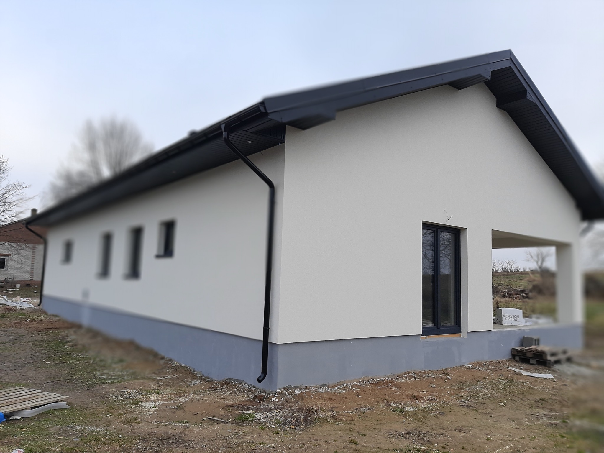 Nowoczesny, parterowy dom z jasną elewacją i ciemnoszarą podmurówką oraz stolarką okienną, widok z zewnątrz na tle pochmurnego nieba i niezagospodarowanego terenu.