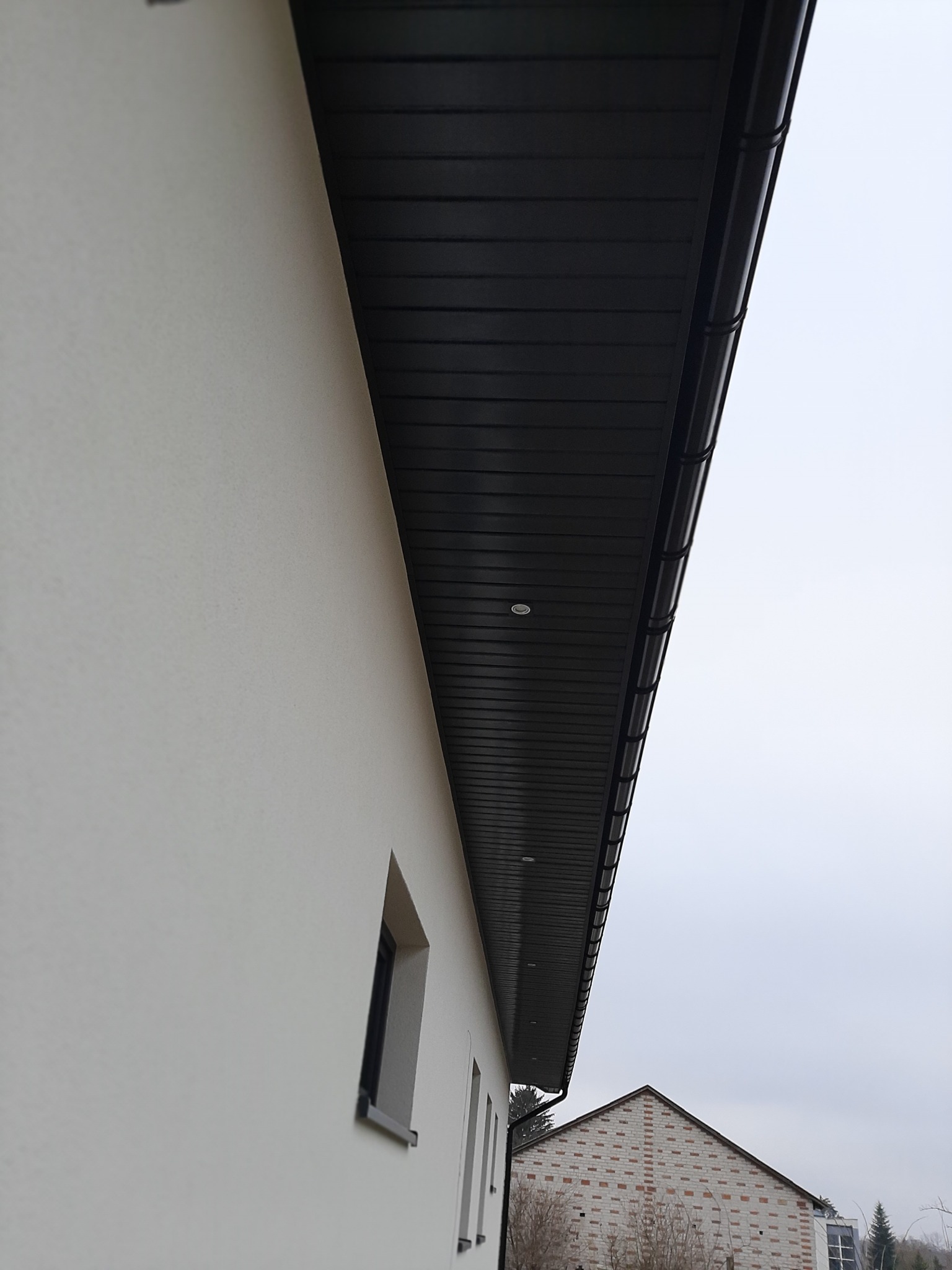 Elewacja budynku z widocznym ociepleniem, ciemną podbitką z oświetleniem punktowym oraz fragmentem okna i dachu innego budynku w tle.