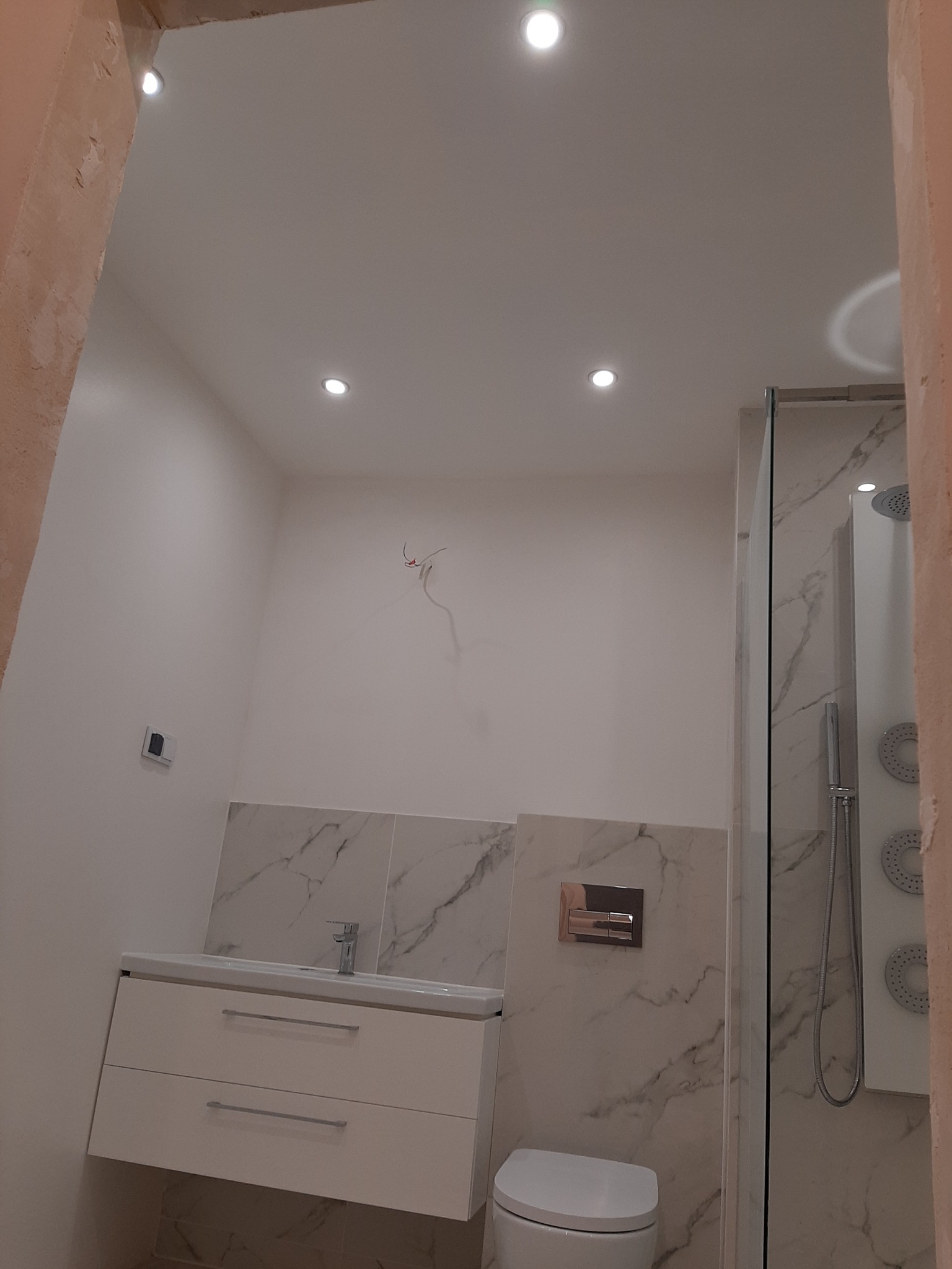 Nowoczesna łazienka w trakcie wykańczania: biała szafka z umywalką, wisząca toaleta, kabina prysznicowa z marmurowym wzorem, surowe przewody elektryczne wystające ze ściany, oświetlenie punktowe...