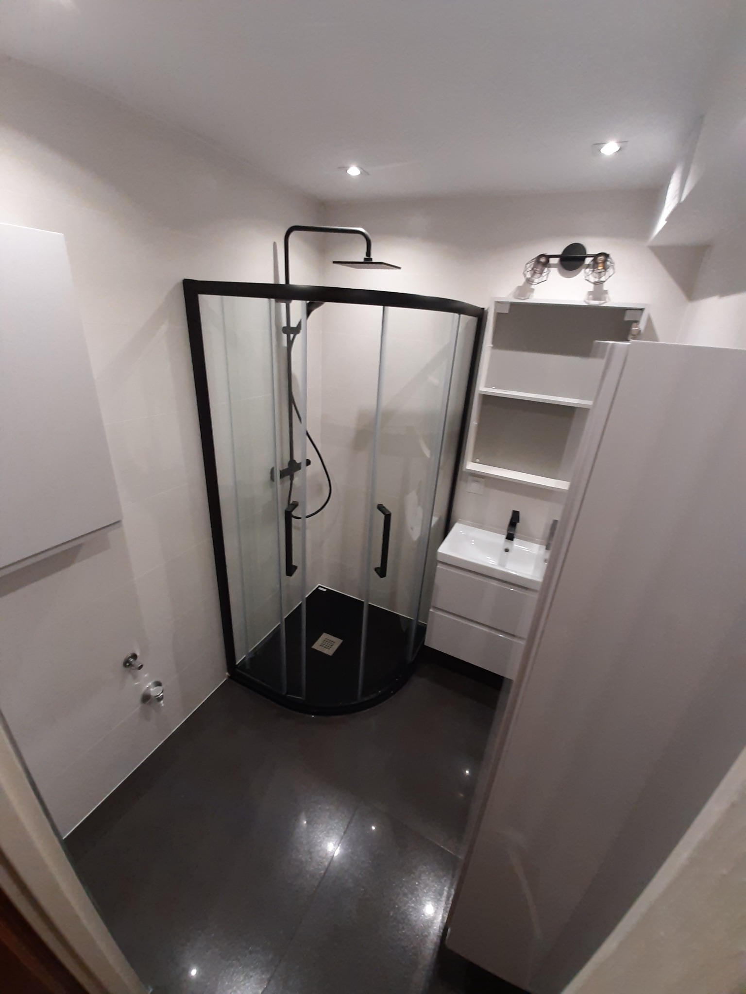 Nowoczesna łazienka z kabiną prysznicową typu walk-in z czarnymi profilami, umywalką w szafce z półkami i ciemną podłogą.