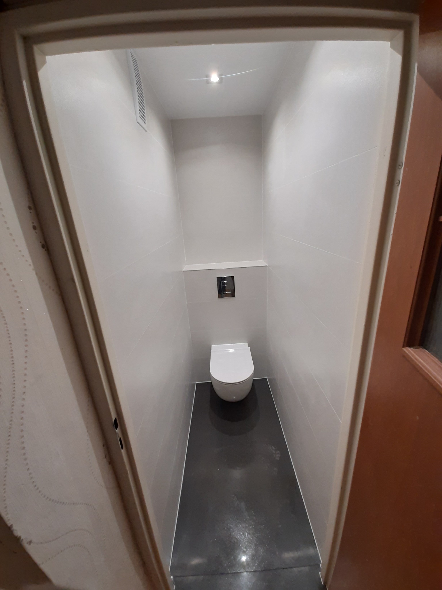 Wąska toaleta w odcieniach szarości i bieli, z podwieszaną muszlą klozetową i czarną podłogą, widok z perspektywy wejścia, z widocznym fragmentem wzorzystej tapety na ścianie po lewej stronie.