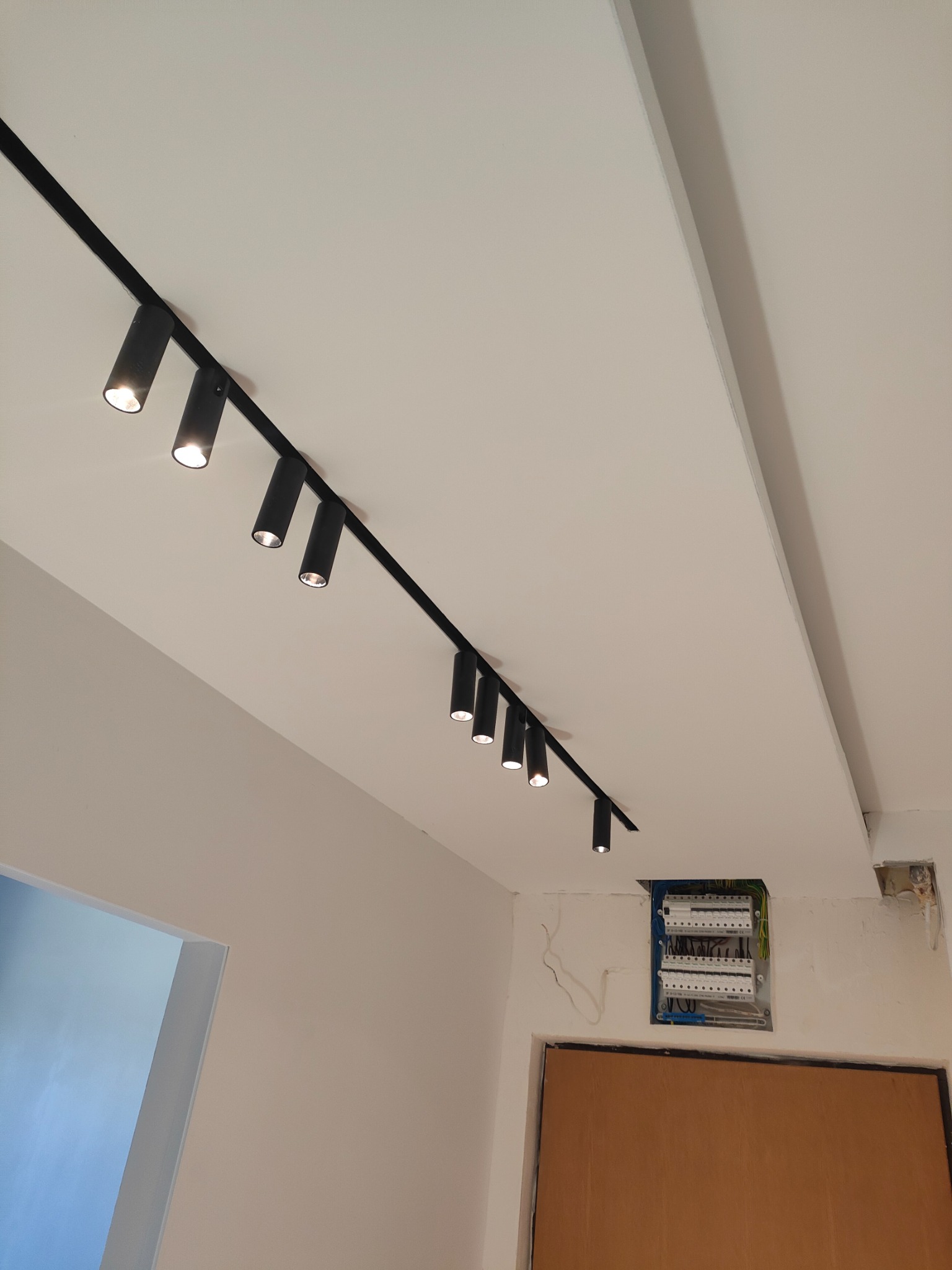 Biały sufit z zamontowanym czarnym systemem szynowym z reflektorami punktowymi, widoczna skrzynka elektryczna i fragment drzwi.