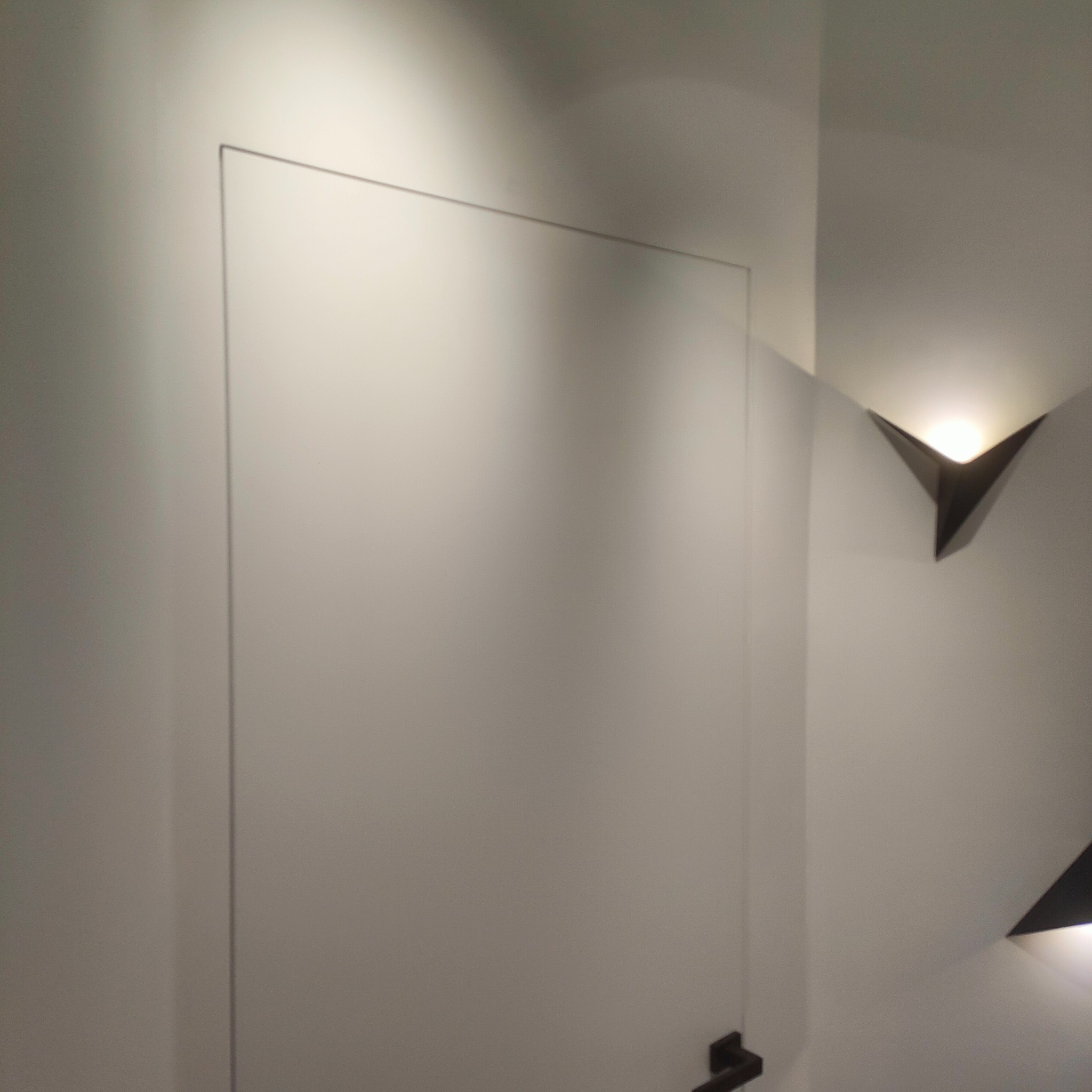 Minimalistyczna, ukryta w ścianie, biała klapa rewizyjna z czarnym uchwytem, oświetlona nowoczesnymi, trójkątnymi lampami ściennymi.