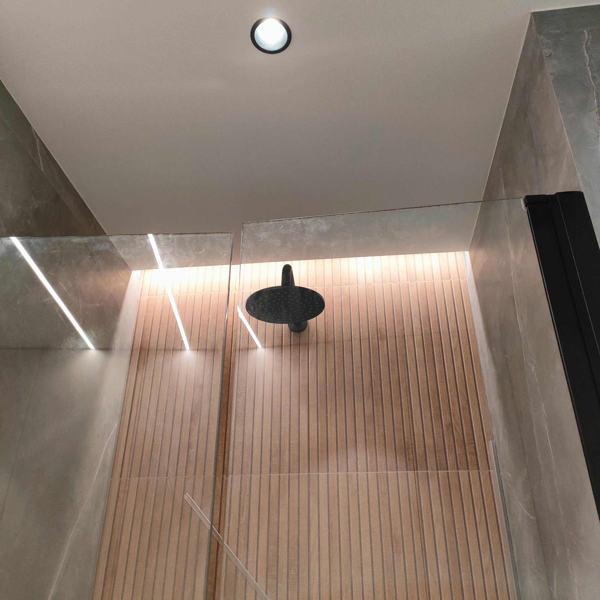 Nowoczesna kabina prysznicowa z deszczownicą, wykończona płytkami imitującymi drewno i szarym kamieniem, oświetlona listwą LED i punktowym światłem sufitowym.